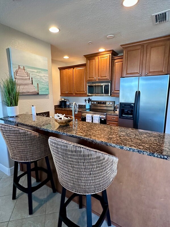 A kitchen with wooden cabinets, a stainless steel refrigerator, a microwave, and a granite countertop. There are two wicker bar stools and a potted plant.
