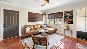 Ein Wohnzimmer mit einem braunen Ledersofa, einem runden Couchtisch, einem Korbstuhl, einem Deckenventilator und einem großen Fenster mit Jalousien.