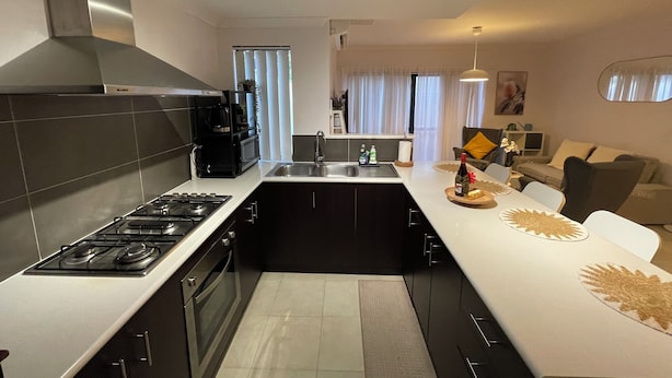 A modern kitchen with a gas stove, black cabinets, and a white countertop. There is a dining area with a sofa and a coffee table. - Cosy White Retreat (Nollamara)