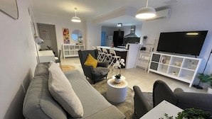 A modern living room with a grey sofa, a coffee table, and a TV. There is a kitchen area in the background with a stove and a dining table. - Cosy White Retreat (Nollamara)
