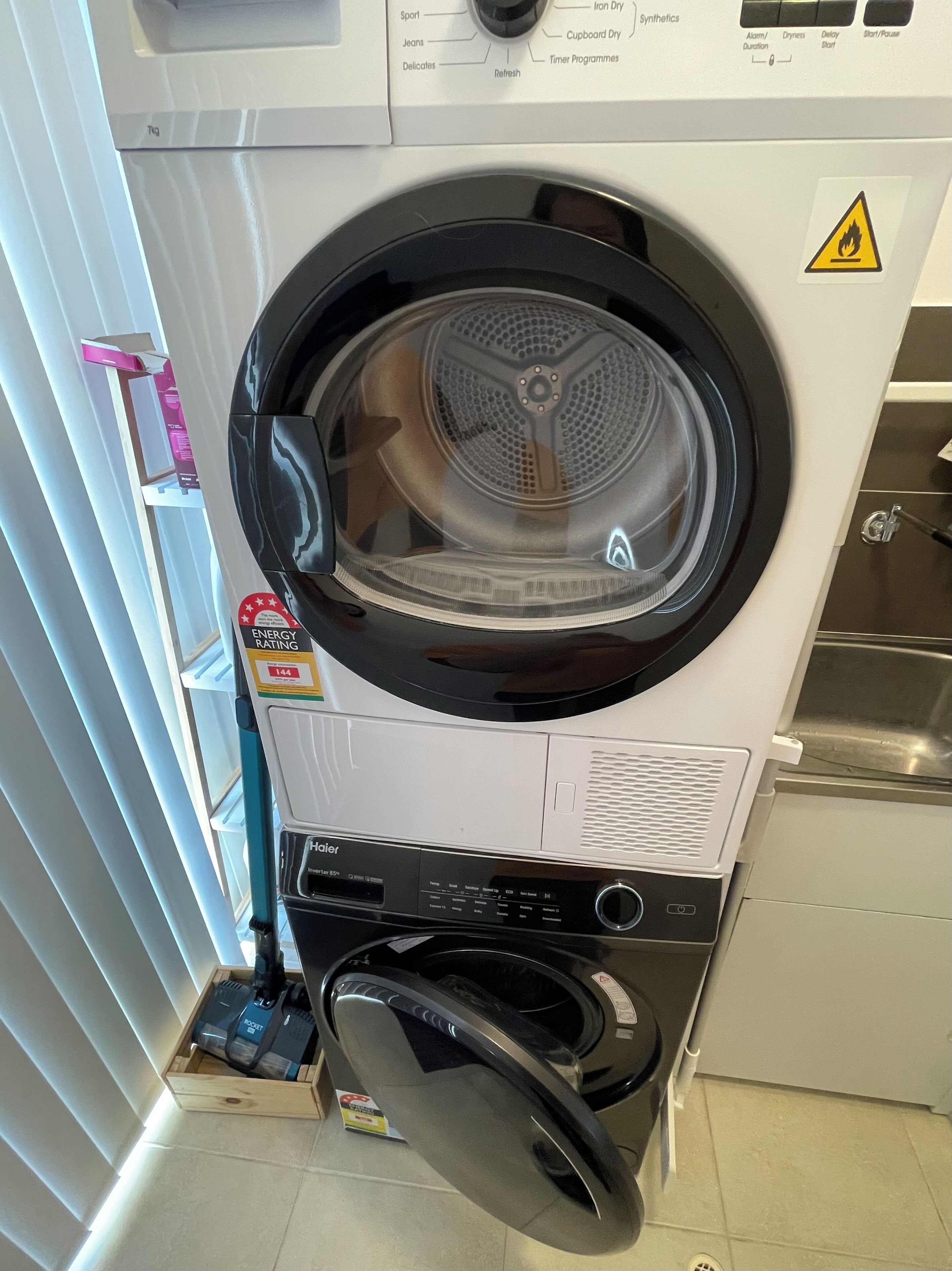 A front-loading washing machine with a control panel, a safety warning label, and a power cord.