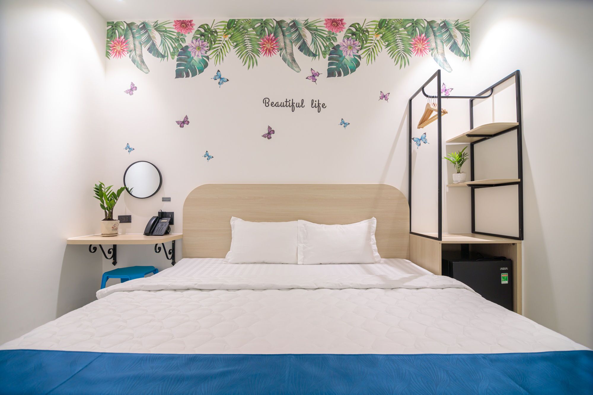 A bedroom with a bed, a bedside table with a phone, a mirror, and a wall-mounted shelf with a plant.