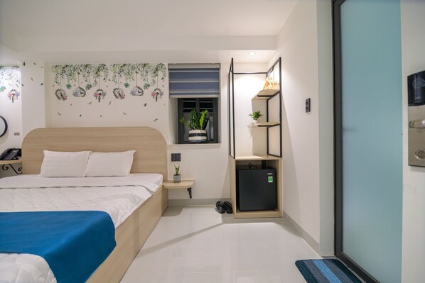 A hotel room with a bed, a small bedside table, a wall-mounted shelf, a potted plant, and a door with a handle.