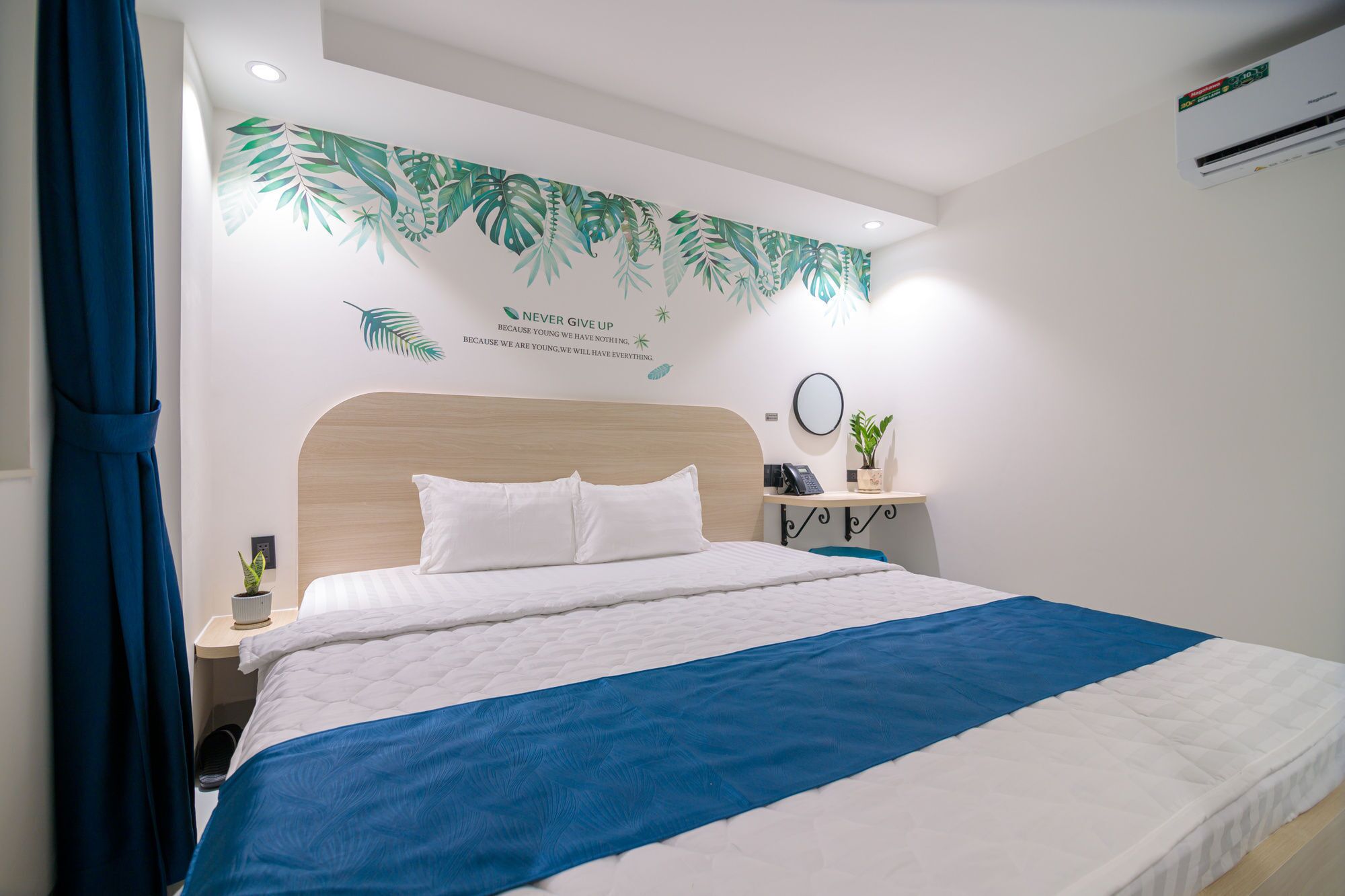 A neatly made bed with a blue and white bedspread, a wooden headboard, a bedside table with a lamp, a wall-mounted air conditioner, and a motivational quote on the wall.