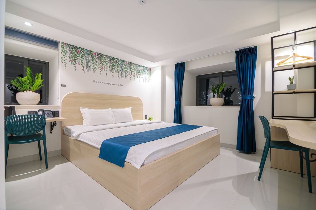 Una habitación de hotel moderna con una cama de madera, un escritorio y sillas. Hay una ventana con cortinas, una planta en el escritorio y una pared con un diseño floral.