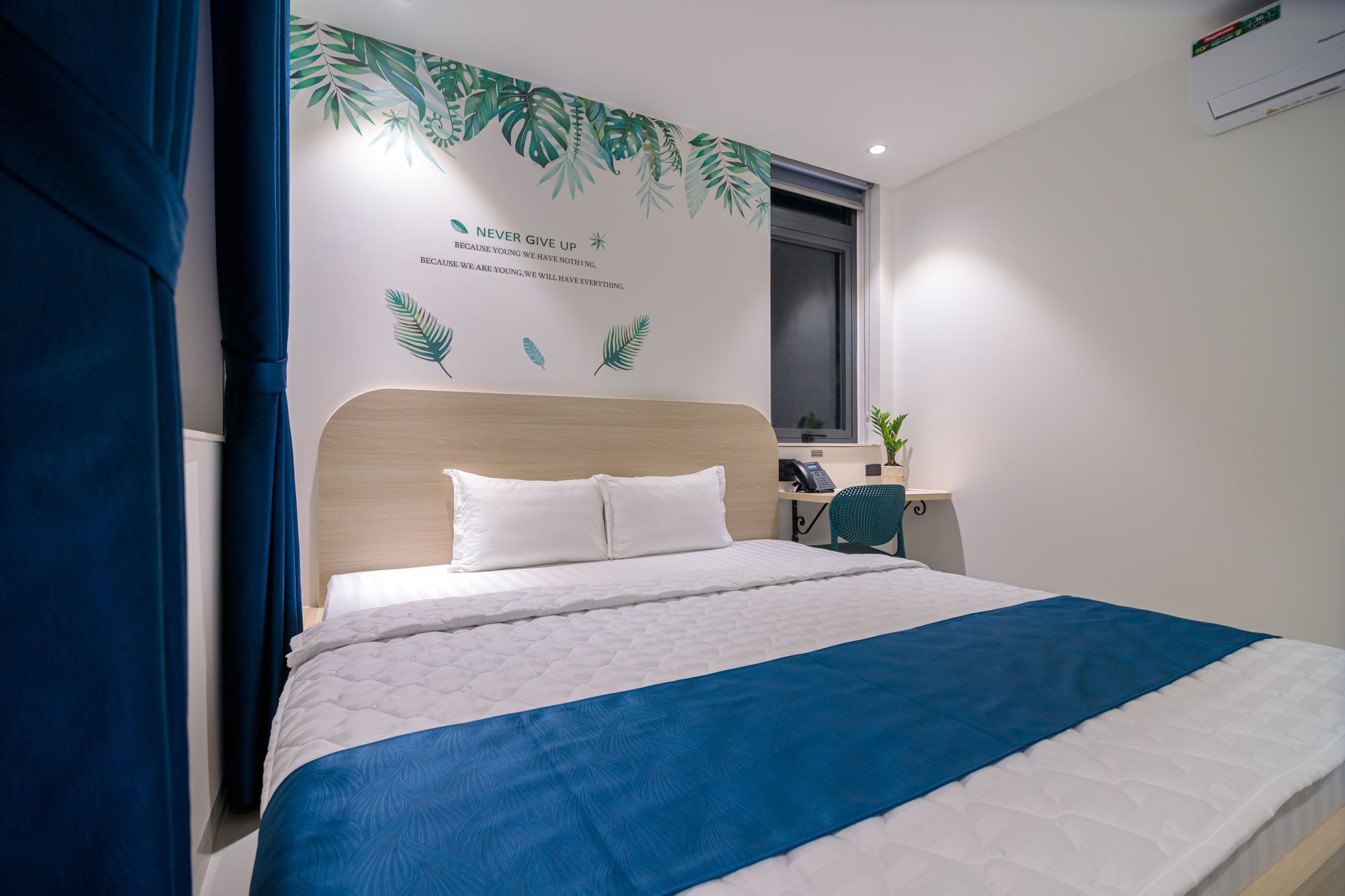 A neatly made bed with a blue and white bedspread, a wooden headboard, and a wall with a motivational quote and leaf decals.