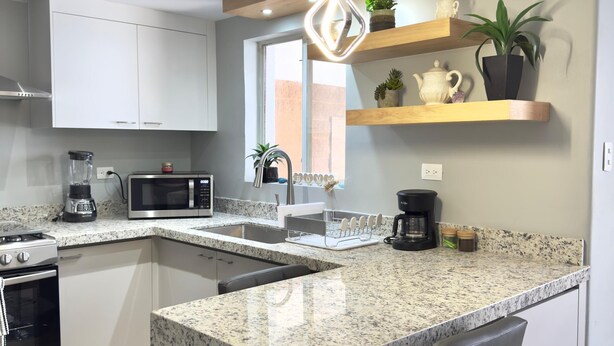 Uma cozinha moderna com bancadas de granito, forno embutido, micro-ondas e máquina de café. Há banquetas de bar e um pendente.