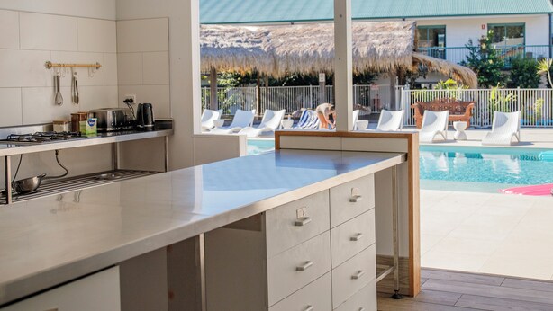 A modern kitchen with a large island, a dining area, and a view of a pool and outdoor seating. - Freedom Backpackers Hostel (Rainbow Beach)