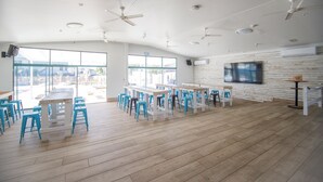 A spacious room with multiple tables and chairs, large windows, ceiling fans, and a mounted television. - Freedom Backpackers Hostel (Rainbow Beach)