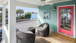 A covered porch with wicker chairs, a ceiling fan, and a view of the water. - Wrightsville Beach Cottage with Stunning Sunset Views, Steps From the Water (Wrightsville Beach)