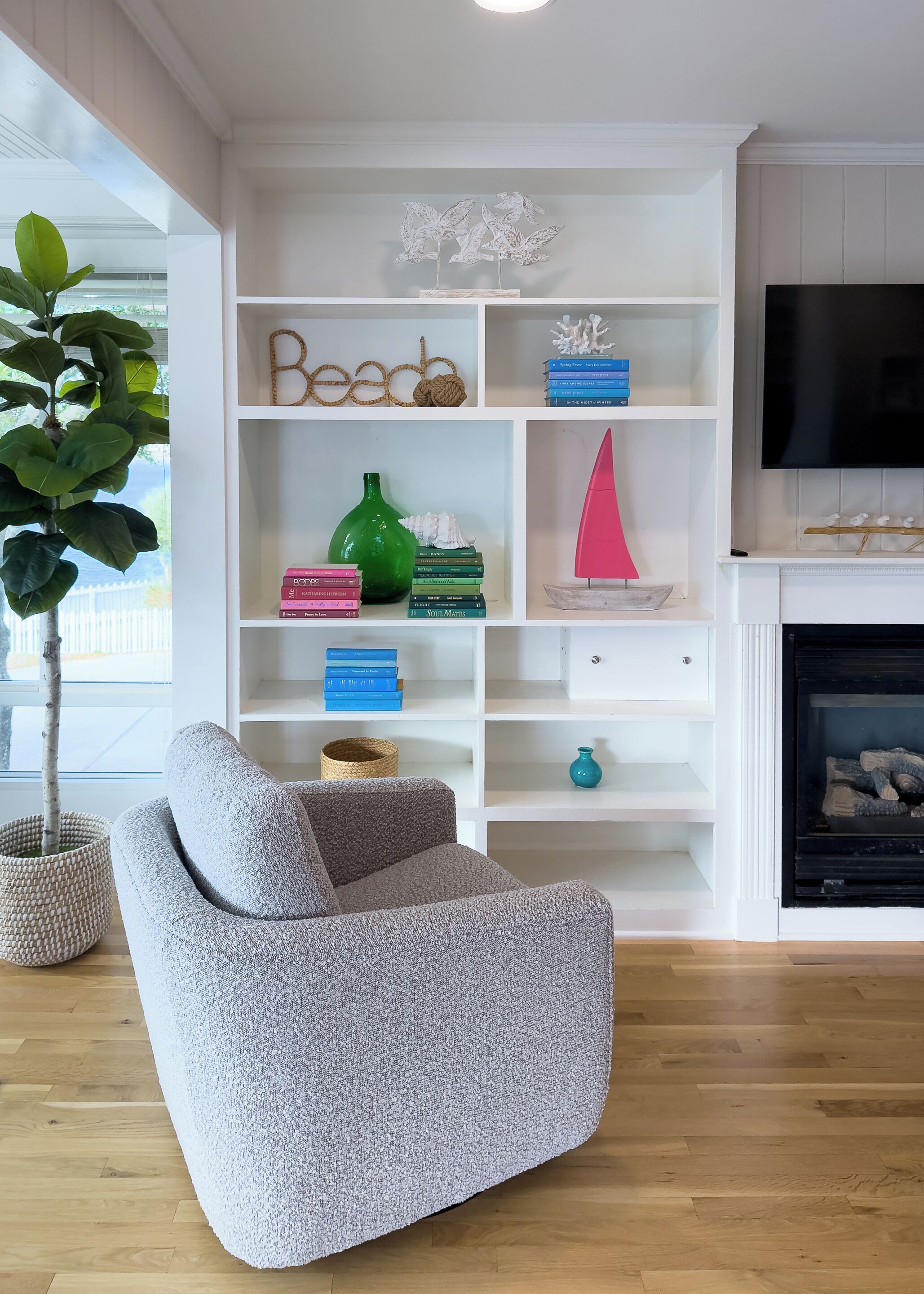 Un salon avec un fauteuil gris, une bibliothèque remplie de livres et d’objets décoratifs, une cheminée et une grande fenêtre avec une plante.