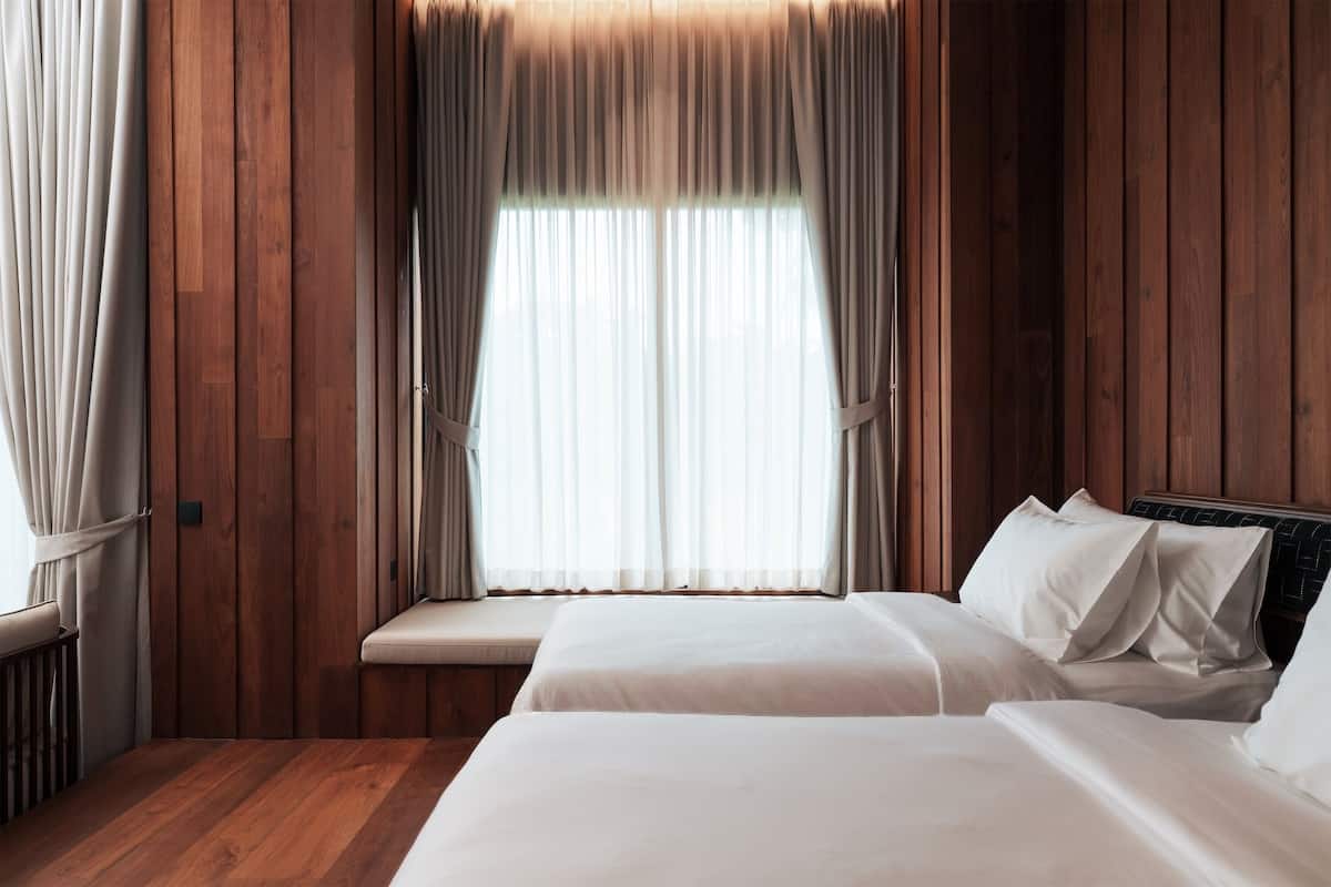 A room with two beds, wooden paneling, and a large window with curtains.
