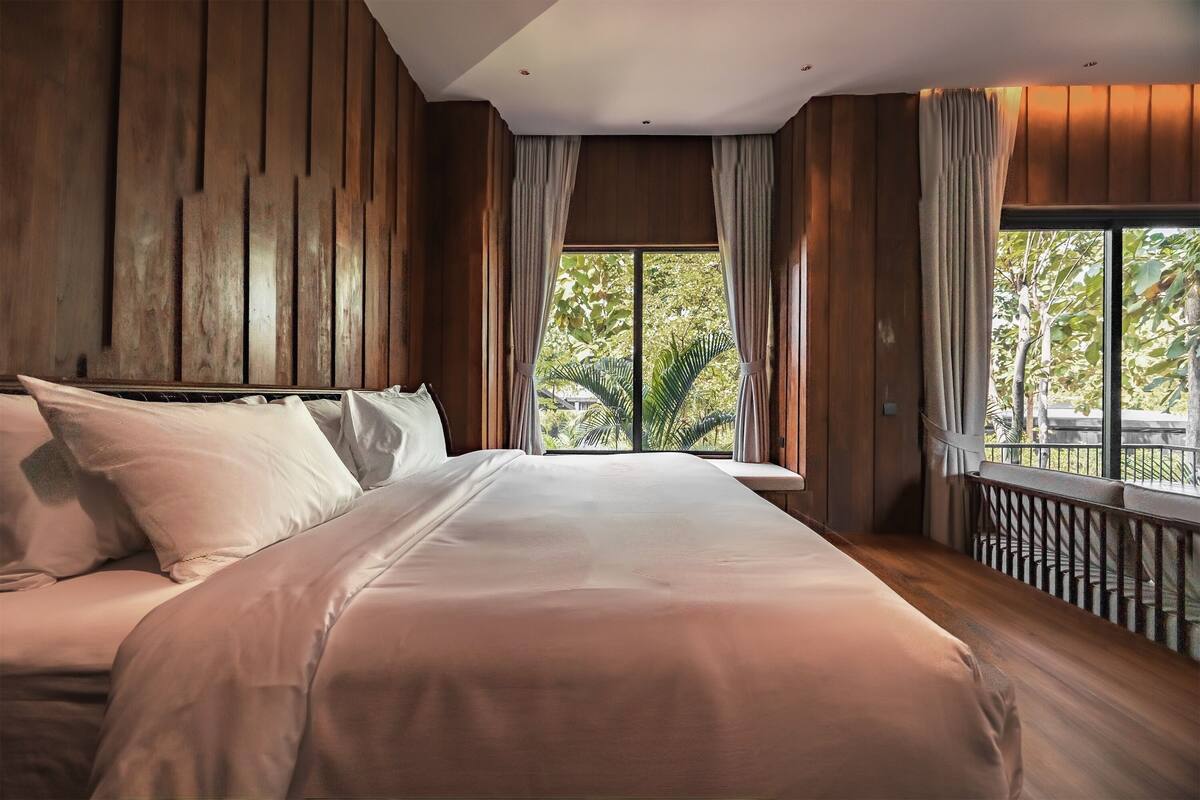 A bedroom with a large bed, wooden panel walls, a window with curtains, and a balcony railing.