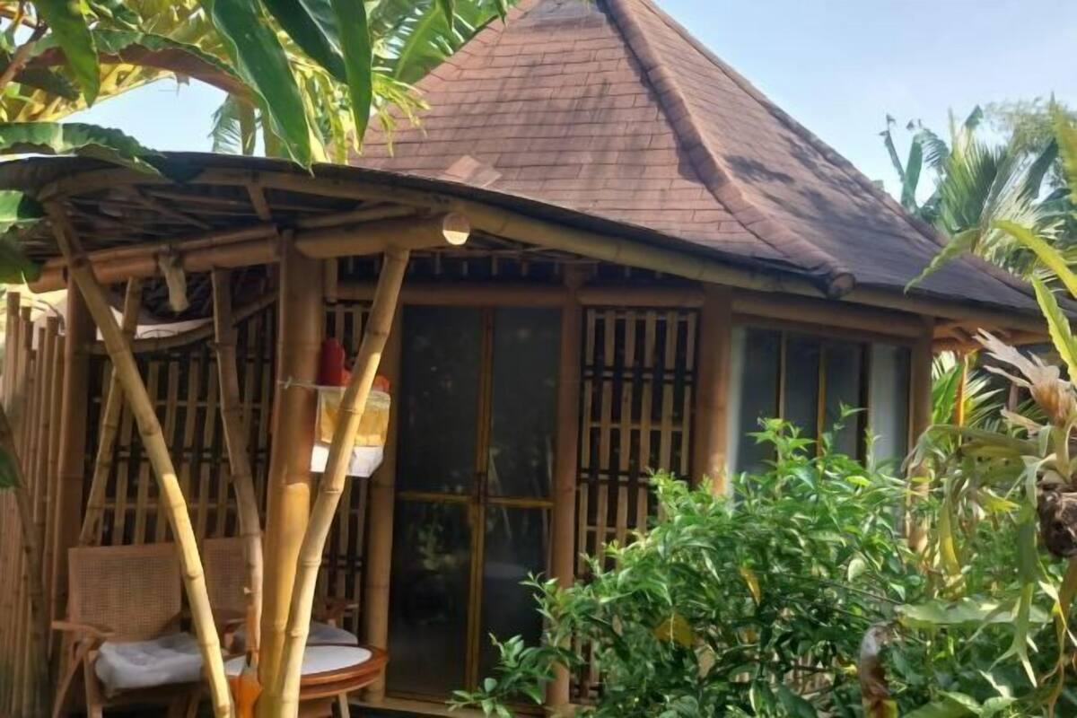 Une structure en bambou surmontée d’un toit de chaume, dotée d’une porte coulissante en verre et d’un petit coin salon avec des coussins.