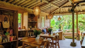 A rustic wooden interior with tables and chairs, a ceiling fan, and a view of lush greenery outside.