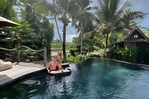 Eine Person in einem kleinen Boot auf einem Pool, umgeben von üppiger Vegetation und einem traditionellen Haus.