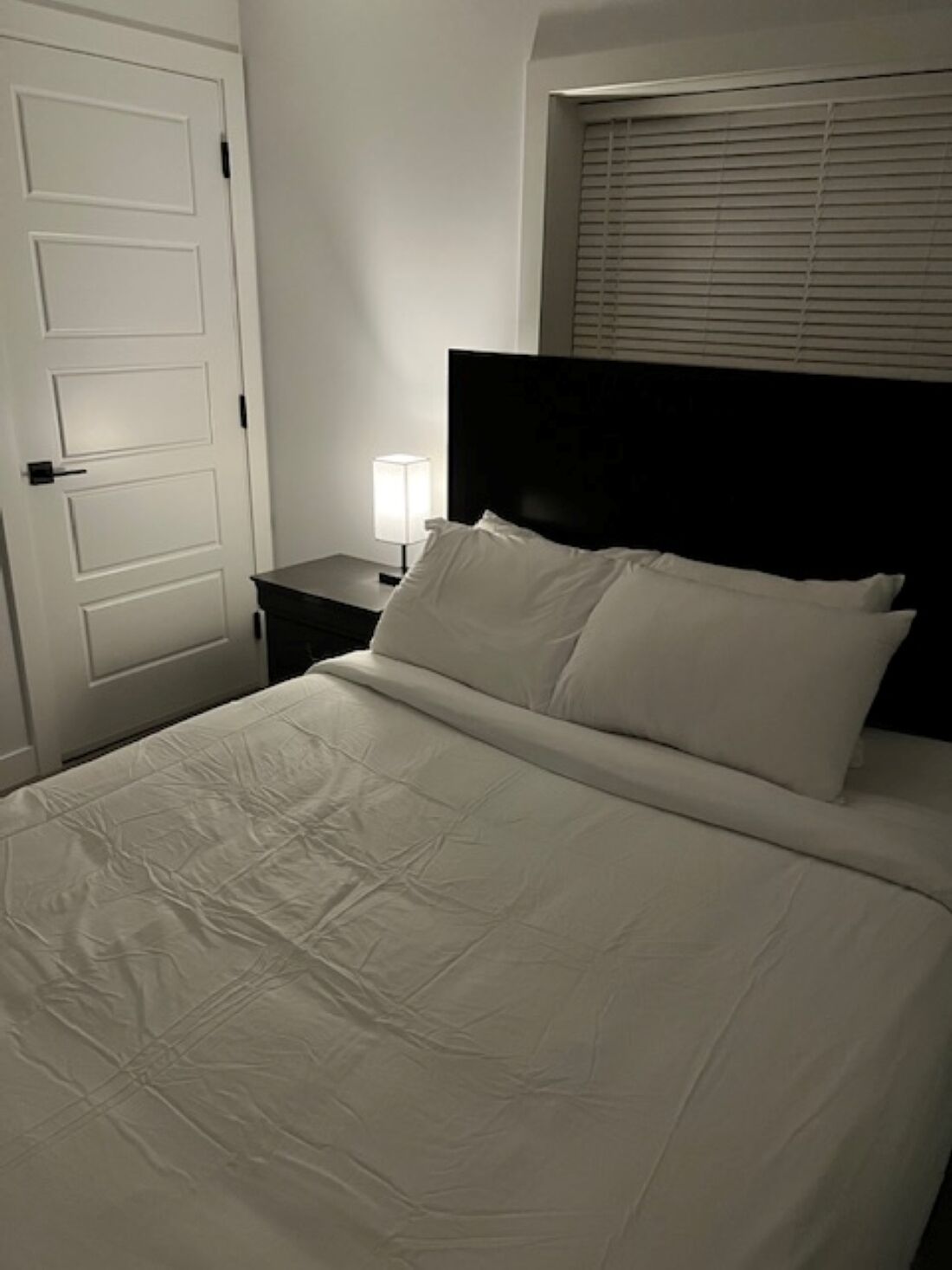 A neatly made bed with white linens, a bedside table with a lamp, and a closed door.