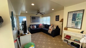 A living room with a dark blue sofa, a ceiling fan, a wall-mounted TV, and a framed poster on the wall. - 3-bdrm in Ship Bottom. 2 blocks from beach and bars. Surfboards included! (Ship Bottom)
