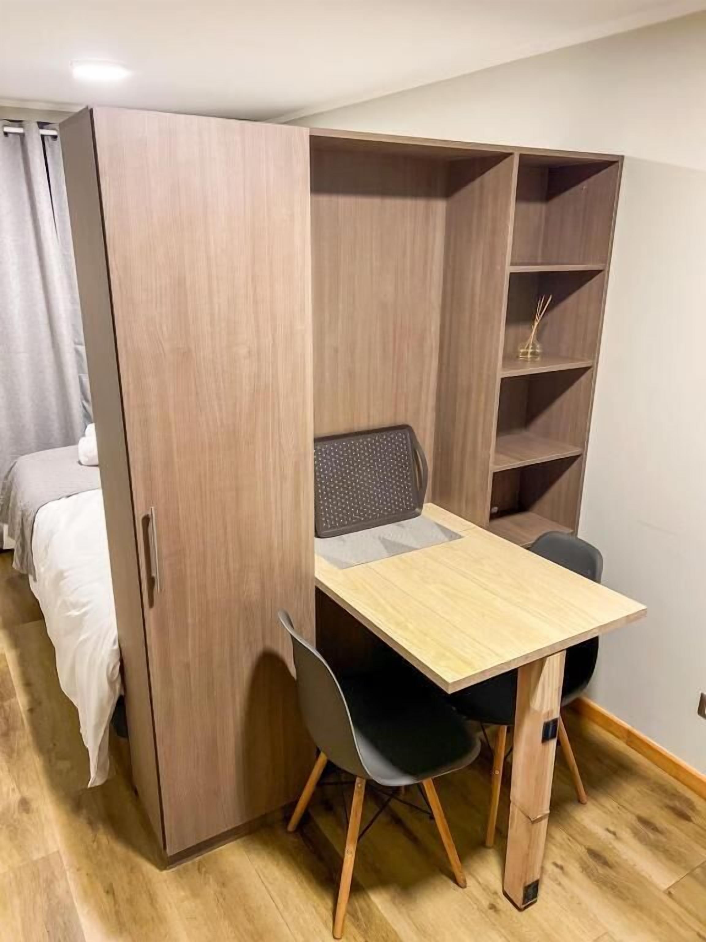 A compact room with a foldable wooden wardrobe, a small desk with a chair, and a bed.