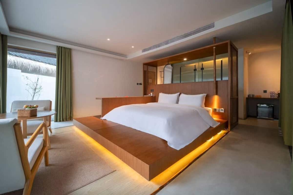 Una habitación de hotel moderna con una cama grande, cabecera de madera, una mesita de noche y un pequeño rincón de descanso con una silla y una mesa.