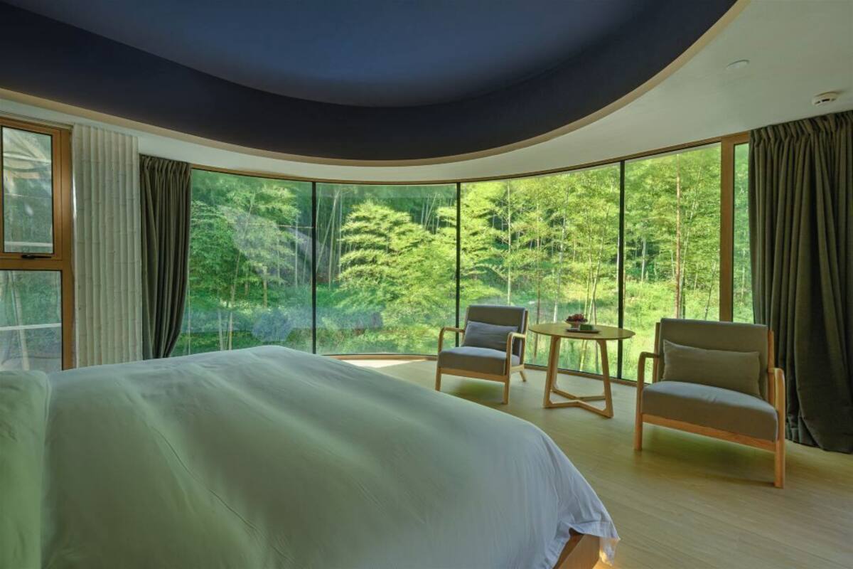 Un dormitorio con una cama grande, dos sillones, una mesita y vistas a un bosque a través de amplias ventanas.