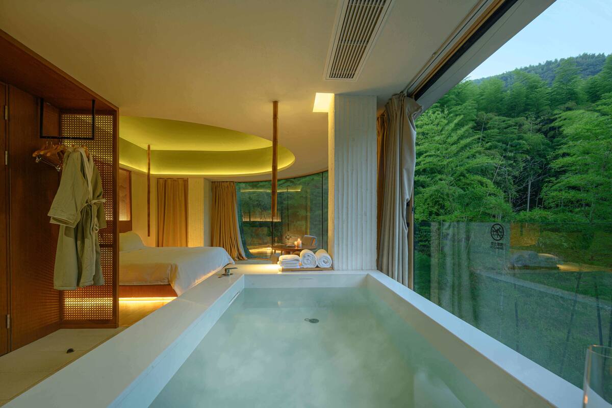 Una habitación de hotel moderna con una bañera grande, una cama y vistas a un área boscosa.