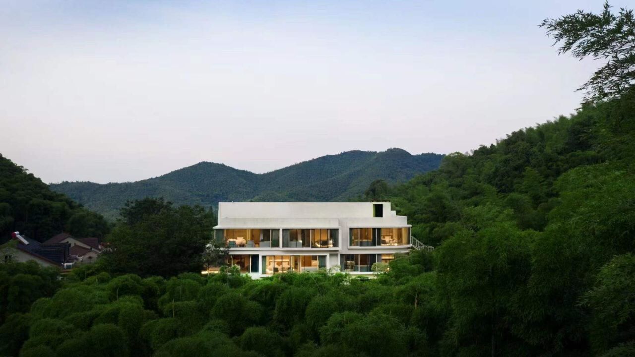 Une maison moderne à deux étages, dotée de grandes fenêtres, entourée d’une végétation luxuriante et avec des collines en arrière-plan.