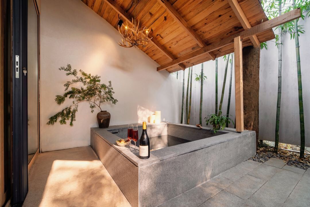 A stone hot tub with a wooden ceiling, surrounded by bamboo plants and a small potted plant.