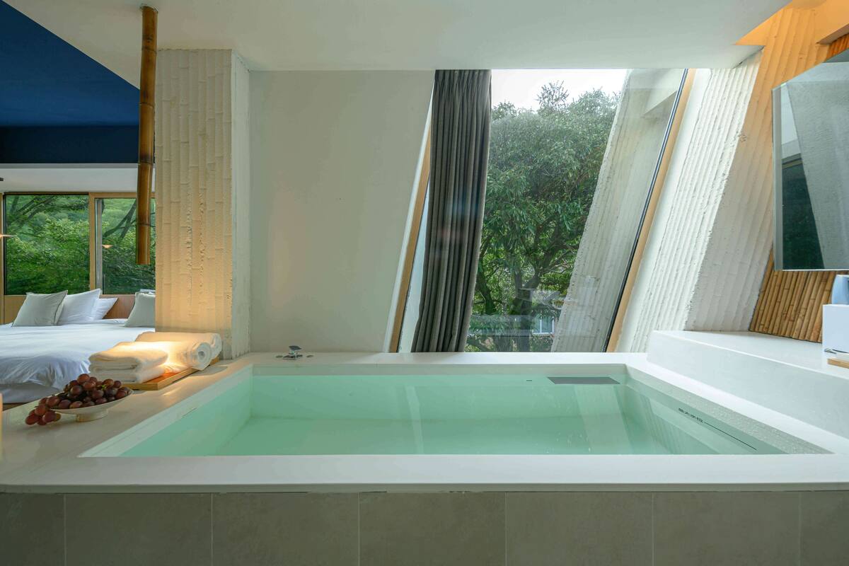 Un baño moderno con una bañera grande, una cama con ropa blanca y vistas a la vegetación.