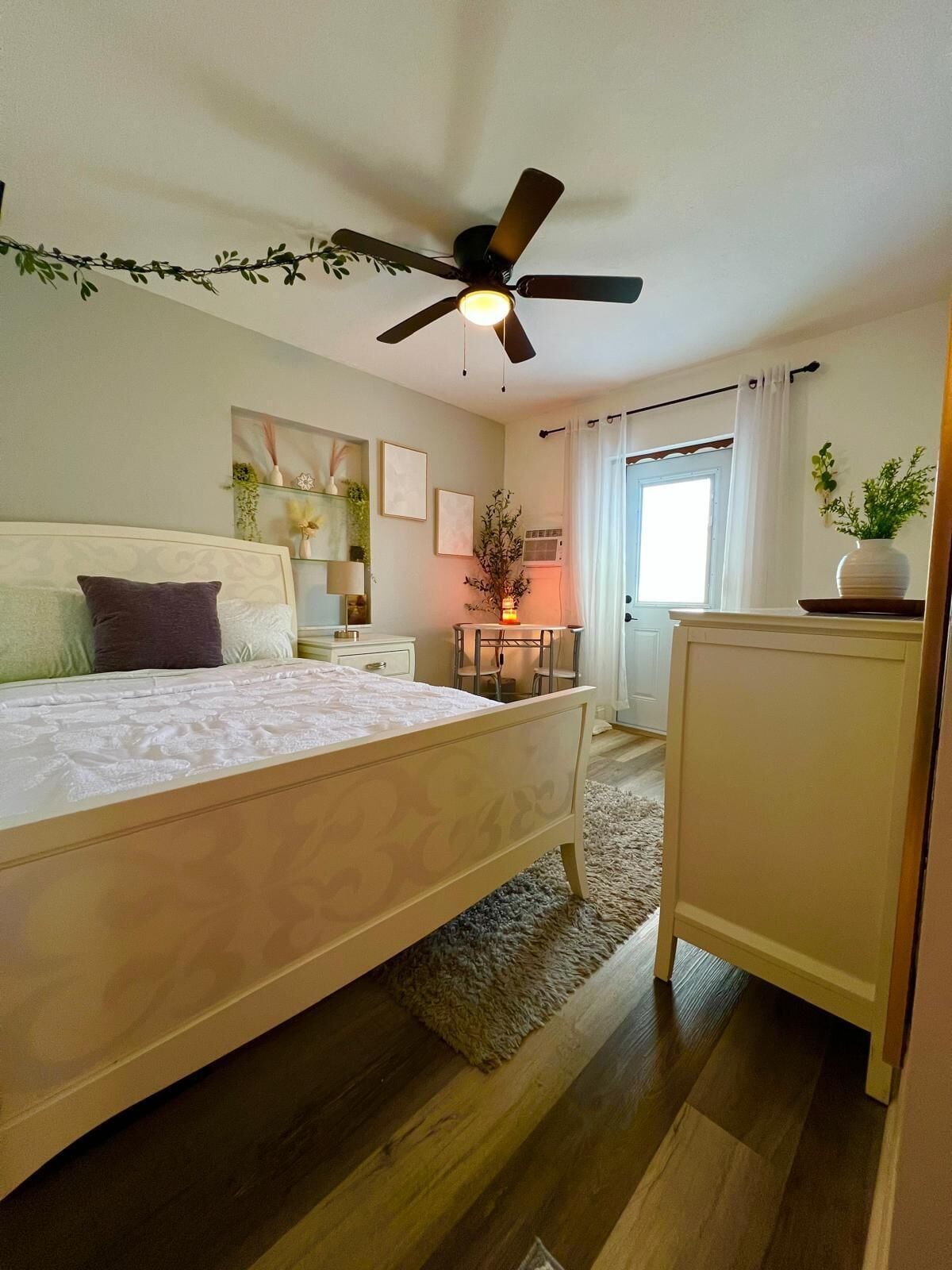Quarto com cama, mesa de cabeceira, cômoda, ventilador de teto, janela com cortinas e uma pequena mesa com uma planta.