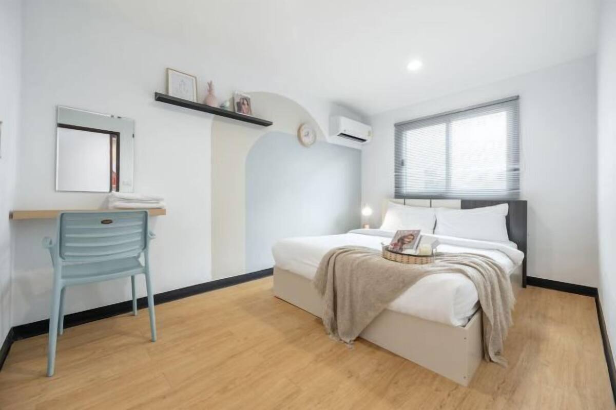 A bedroom with a bed, a desk, a chair, a shelf with items, and a window.