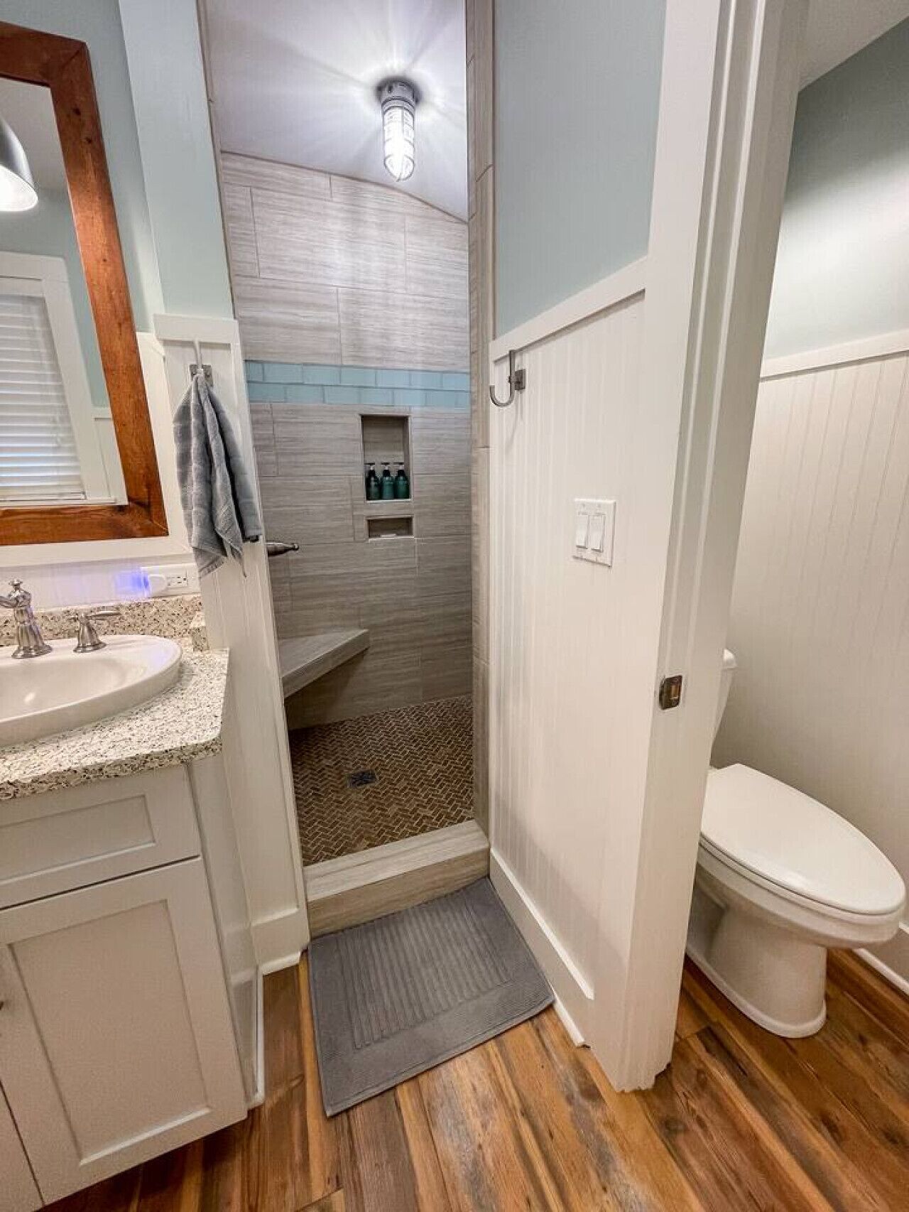 A bathroom with a wooden floor, a white sink with a granite countertop, a shower with a tiled wall, and a white toilet.