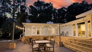 An outdoor patio with wooden furniture and string lights, surrounded by a white building with large windows. - Retreat for families, walk to beach, stocked kitchen, pet friendly, fenced yard (Pawleys Island)