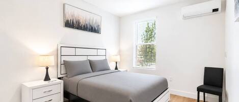 Un dormitorio moderno con cama, mesitas de noche, una silla, ventana con persianas y aire acondicionado.