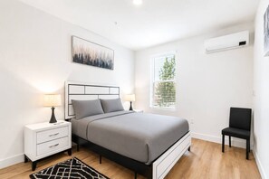 Ein modernes Schlafzimmer mit Bett, Nachttischen, einem Sessel, einem Fenster mit Jalousien und einer Klimaanlage.
