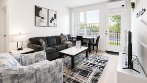 A modern living room with a sofa, a coffee table, and a TV. There is a dining area with a table and chairs. The room has large windows with blinds, and a door leading outside.