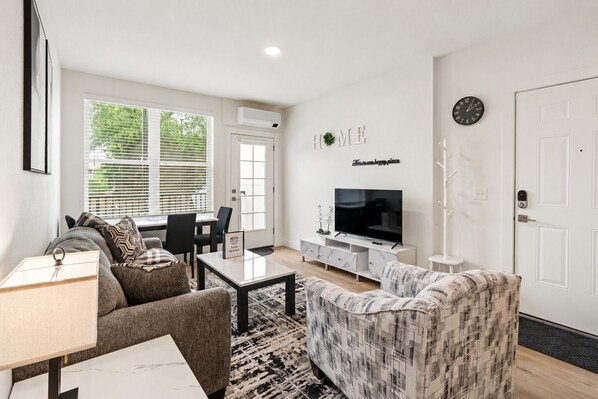 A modern living room with a sofa, armchair, TV, and a dining area with chairs and a table.