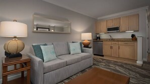 A modern living room with a grey sofa, two lamps, a coffee table, and a kitchen area with wooden cabinets and a microwave.