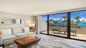 A hotel room with a sofa, a coffee table, a lamp, a nightstand, and a large window with a view of a beach and palm trees.