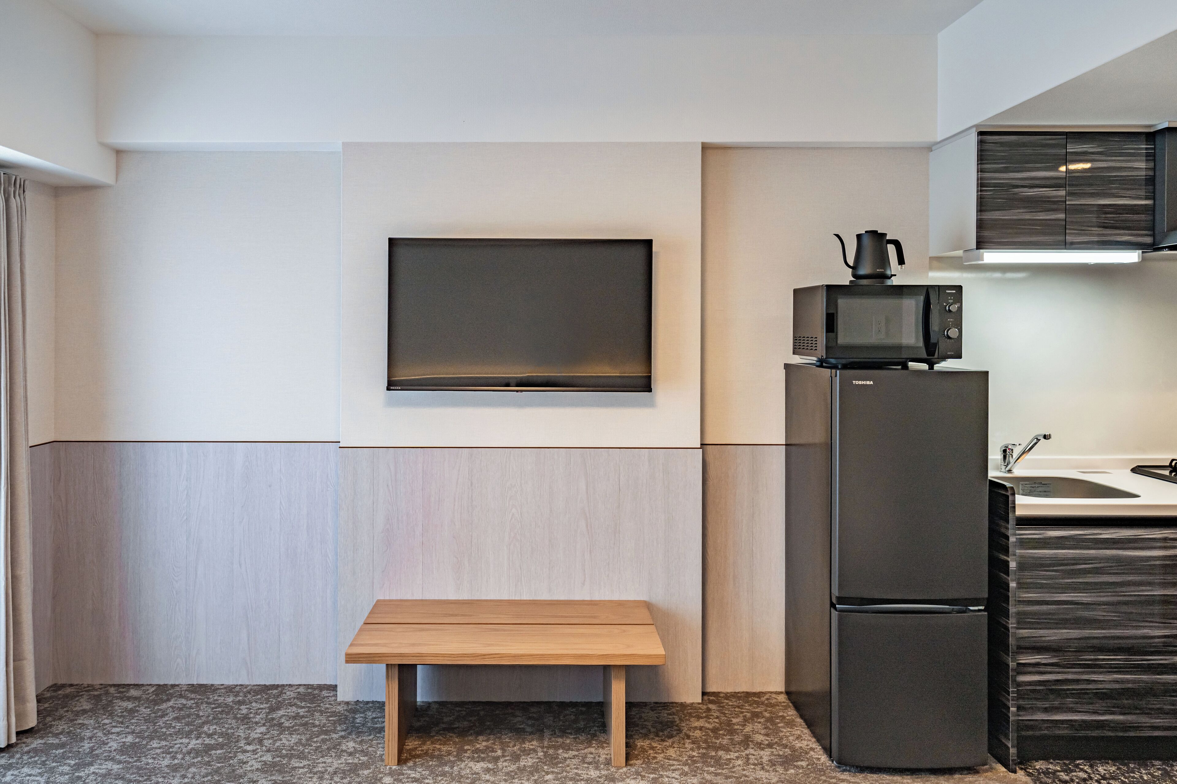 A compact kitchenette with a flat-screen TV mounted on the wall, a microwave, and a refrigerator.