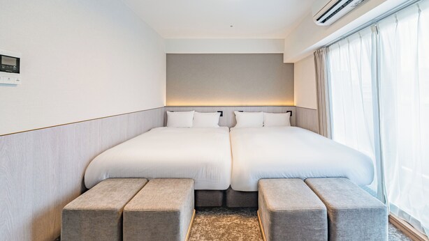 A hotel room with a double bed, two bedside tables, a wall-mounted air conditioner, and a large window with curtains. - Apartment Hotel 11 Dotonbori 7 (Osaka)