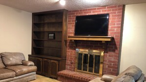 Un salon doté d’une cheminée en briques, d’un téléviseur à écran plat fixé au-dessus, d’un grand meuble de rangement en bois et de deux canapés en cuir.