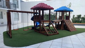 A wooden play structure with a swing set and climbing wall, surrounded by a green artificial turf area.