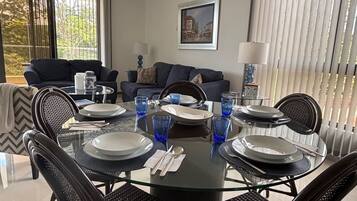 Un comedor con una mesa de vidrio preparada para cuatro personas, rodeada de sillas negras. Se ve un sofá y una lámpara al fondo.
