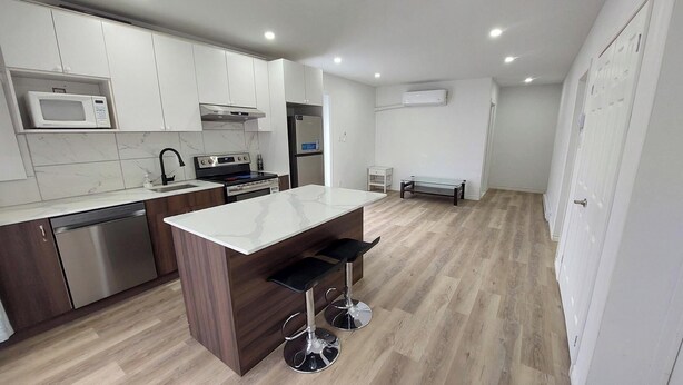 A modern kitchen with white cabinets, a stainless steel dishwasher, a microwave, a stove, and a sink. There is a wooden island with two bar stools. - Bright 2BR Near River · Private Parking (McMasterville)