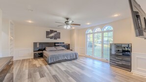 Une chambre à coucher comprenant un lit, des tables de chevet, un ventilateur de plafond, une grande fenêtre et un téléviseur intégré.