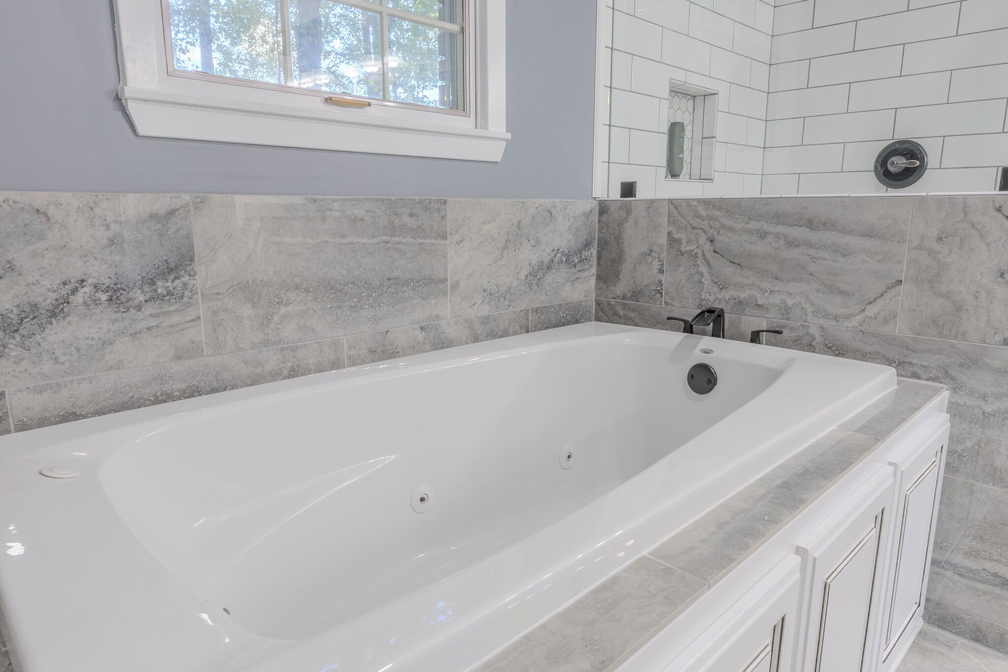 A modern bathroom with a large white bathtub, marble tiles, and a window.
