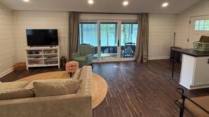 A living room with a television, a sofa, a chair, and a view through a sliding glass door to an outdoor area. - Waterfront Paddles & Pours Cottage on Claytor Lake (Hiwassee)