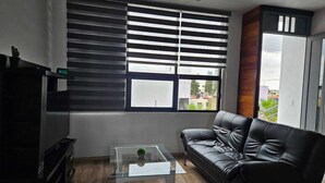 A modern living room with a black leather sofa, a glass coffee table, and a large window with blinds. - Mia La Perla Apartment, very close to La Perla District, Plaza del Sol, Expo GDL (Zapopan)