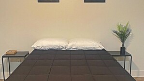 A neatly made bed with a dark quilt, two bedside tables with lamps, and a small potted plant on the right. - Deep Ellum-Sleeps 5-Balcony-Pool-Medical District (Dallas)